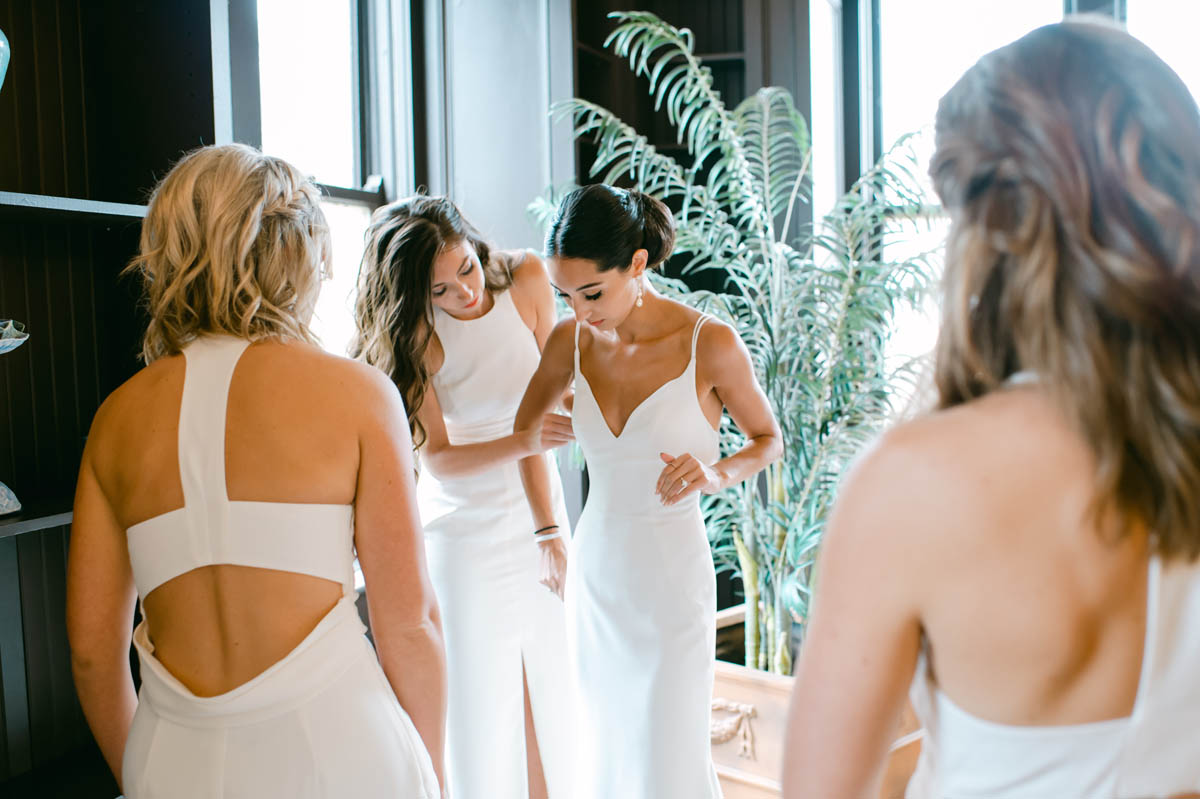 Bridesmaids in white dresses helping bride get dressed.