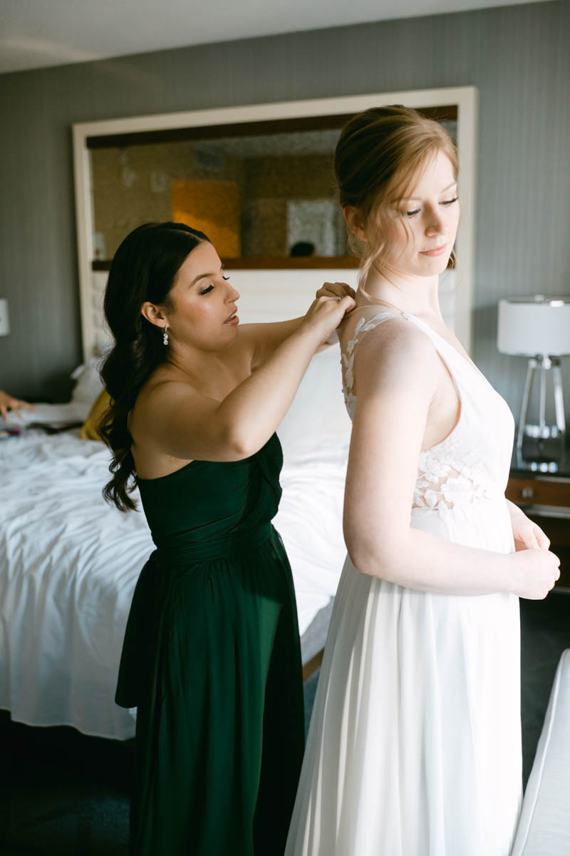 Bridesmaid in green dress helping bride into her dress.