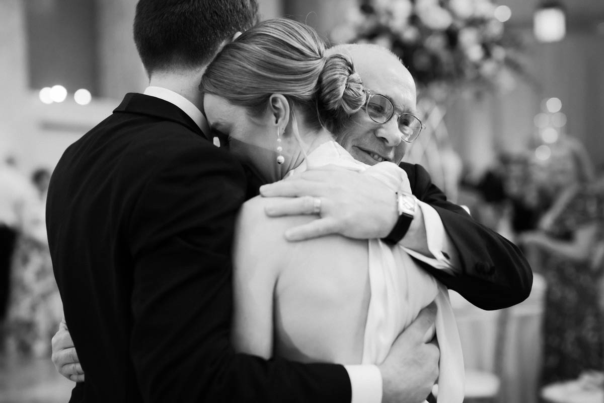Black and white portrait of bride and groom embracing dad after toasts